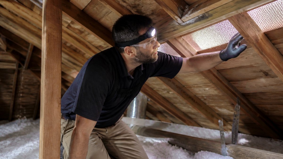 Homeowner wearing safety gear inspecting roof rafters and soffit vents to troubleshoot attic ventilation issues.